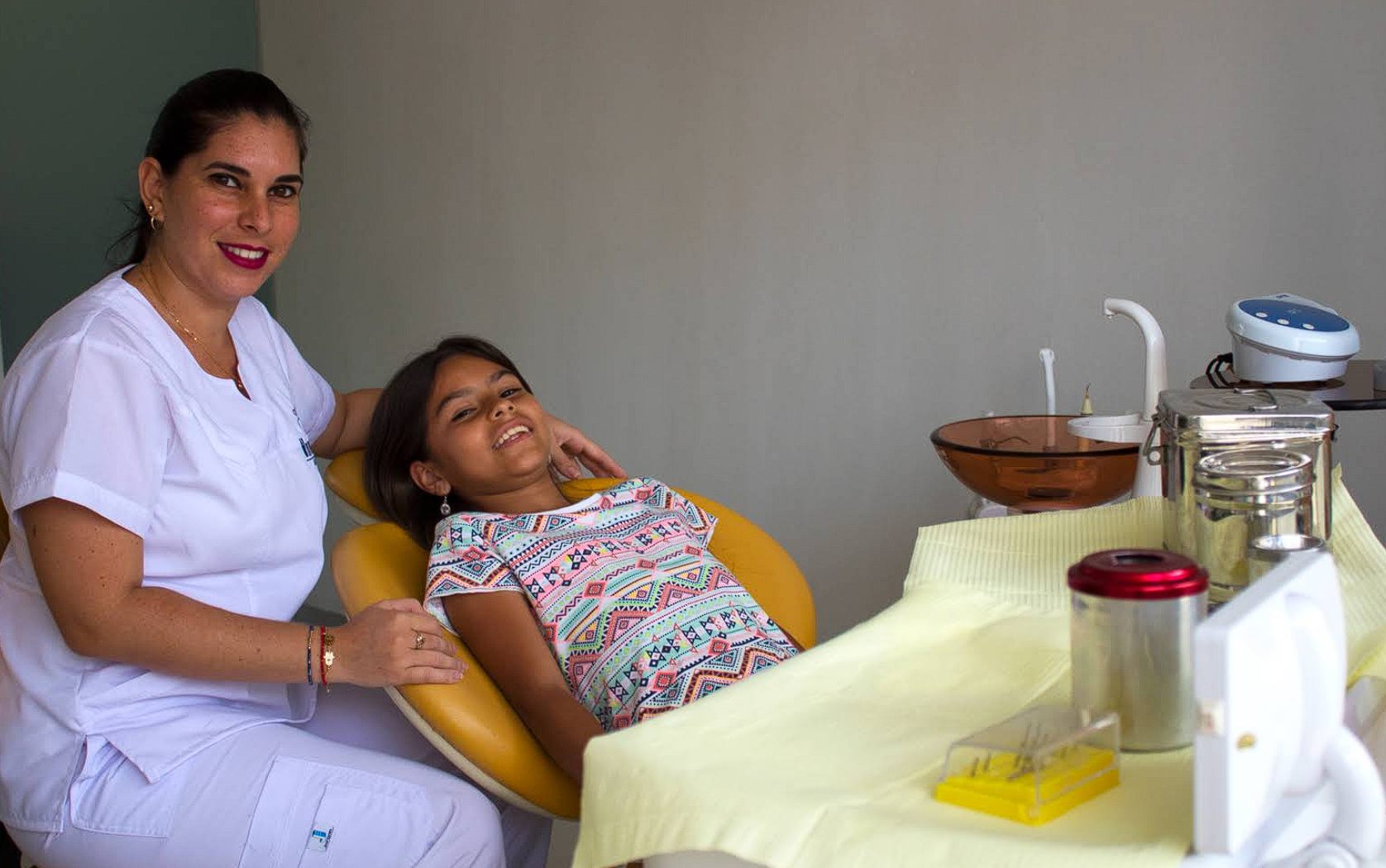 Odontóloga sonriendo junto a una niña sentada en el sillón dental amarillo